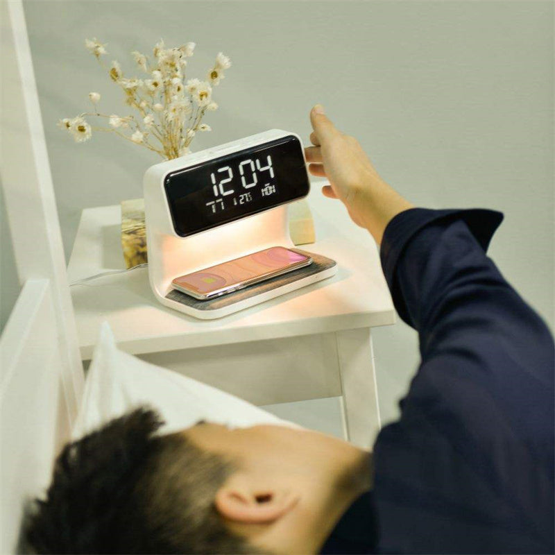 Person using a digital alarm clock on a nightstand with a smartphone on a stand.