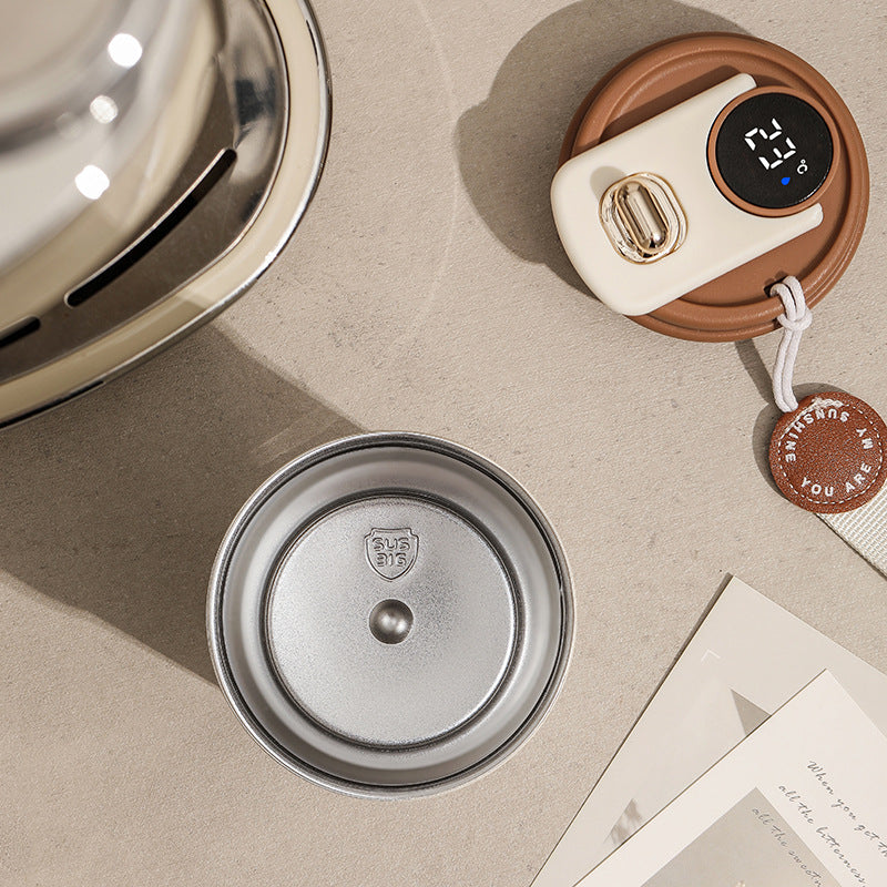 Metallic lid, white coffee electronic device with digital display, and brown leather case on a beige surface.